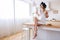 Positive cheerful young woman on table in kitchen. Carefree housekeeper holding cup and croissant in hands. Smile. Alone