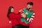 Positive couple dressed in Christmas funny sweaters. Woman tickling her boyfriend over red background.