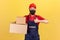 Positive courier in uniform and hygienic mask holding cardboard parcels and showing thumbs up, satisfied with fast service,