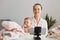 Positive delighted young female blogger broadcasting livestream at home, together with her infant daughter, filming content for