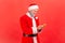 Positive elderly man with gray beard wearing santa claus costume holding cellphone in hands, scrolling web pages, ordering