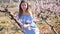 Positive european girl in blue dress standing under blossom peach tree at sunny spring day