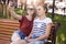 Positive female teenagers sit on wooden in bench in green park, pose for making selfie, look at smart phone, smile positively, do