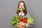 Positive funny smiling woman embracing fresh vegetables returns from supermarket wearing striped shirt  over gray