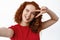 Positive ginger girl with curly hairstyle, showing tongue and v-sign over eye, taking selfie or record video on