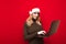 Positive girl in santa christmas hat uses laptop on red background, looks into computer screen and smiles. Isolated. Working at