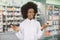 Positive joyful young African American woman pharmacist or doctor, holding in hands two bottles with medicines, standing