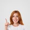 Positive lovely female with shining smile and red hair, points upwards with index finger, glad to present something, dressed in ca
