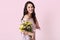Positive optimistic brunette young lady recieves flowers on birthday, wears polka dot stylish dress, smiles gently, poses against