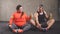 Positive overweight woman and fit guy doing exercises while sitting on the floor.