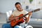 Positive paraplegic black man in wheelchair playing guitar, enjoying music at home