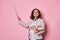 Positive pregnant female teacher in glasses, holding textbooks, smiling at camera, pointing with pointer at copy space