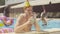 Positive redhead Caucasian man in party hat drinking cocktail in pool and showing thumb up. Portrait of young cheerful