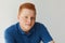 A positive redhead guy with freckles and brown eyes dressed in blue shirt looking mysteriously into camera isolated over white bac