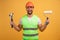 Positive repairman holds hammer and paint roller, wears apron and hardhat, has many building tools, ready for house renovation.