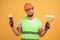 Positive repairman holds paint roller and brush can`t make a choice,wears orange protective construction helmet, reflective