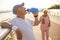 Positive senior man drinks water leaning onto railing while lady runs along footbridge