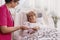 Positive senior patient lying in hospital bed with helpful nurse in pink uniform at her site