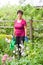 Positive smiling elderly female taking care of plants in summer garden, watering flowers with a watering can