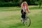 Positive smiling woman dressed in casual clothes, poses on bicycle, has active rest in park, rides on green lawn during sunny