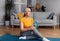 Positive sporty aged lady sitting on fitness mat with bottle of water, having break while exercising at home