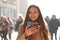 Positive surprised young woman receives good news on her phone while walking in the street with blurred crowd of people on the