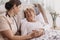 Positive woman lying in bed, helpful doctor in beige uniform supporting her