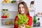 Positive young attractive woman holds lettuce, being vegeterian, stands against opened refrigerator full of fruit and vegetables,