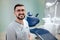 Positive young bearded dentist sit in dentistry room and pose on camera. He wear white robe and glasses.