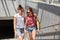 Positive young girls lesbians hold hands, carry rucksack, casual t shirt and shorts, walk near underground have fun together talk