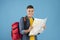 Positive young hiker with backpack and map planning his weekend camping trip route on blue studio background