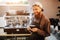 Positive young man stand at coffee machine and smile on camera. He holds balck cup in hands. Guy stand in kitchen.