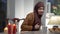 Positive young woman in hijab entering room bringing sweet buns and tea cup sitting down at table. Portrait of confident