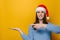 Positive young woman keeps palm raised, points with forefinger against yellow wall for advertisement, dressed in Christmas red hat