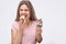 Positive young woman smile on camera. She bite piece of eclair. Model hold cake in another hand. Isolated on grey