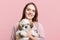 Positve female with long hair and pleased expression holds her favourite little puppy, isolated over pink background. Happy woman