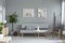 Posters above grey sofa in minimal living room interior with plants and armchair. Real photo