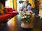 A pot put on wooden table in yellow room. Flowers color are violet, purple, and white with green leaves. Red sofa in the room. No