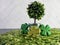 Potted tree sitting on a bed of green leaves with 3 four leaf clovers covered in glitter, against a herringbone tile for St.