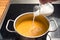 Pouring coconut milk or cream in a vegetable soup from red kuri squash or Hokkaido pumpkin in a steel pot on a black stove,