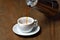 Pouring coffee into empty white porcelain mug with saucer from moka pot coffee maker. Mug on wooden table and stream of coffee