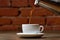 Pouring coffee into white porcelain mug with saucer from moka pot coffee maker. Mug on wooden table and stream of coffee.