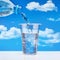 Pouring drinking water from plastic bottle into glass, against blue sky with clouds.