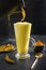 Pouring glass of ayurvedic golden turmeric latte milk with curcuma powder on black. Vertical shot