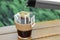 Pouring hot water into glass with drip coffee bag from thermos on wooden table, closeup