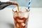 Pouring refreshing soda drink into glass with ice cubes on blurred background