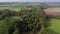 POV from drone flight backwards over a small forest and fields with ponds, a village and wind turbines in the background