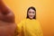 POV of happy smiling influencer wearing yellow sweater taking selfie with camera while on orange background.