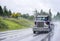 Powerful classic big rig blue semi truck with flat bed semi trailer running on the wet raining road in front of another traffic