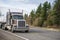 Powerful classic big rig brown semi truck with vertical pipes transporting packed cargo on flat bed semi trailer running on the